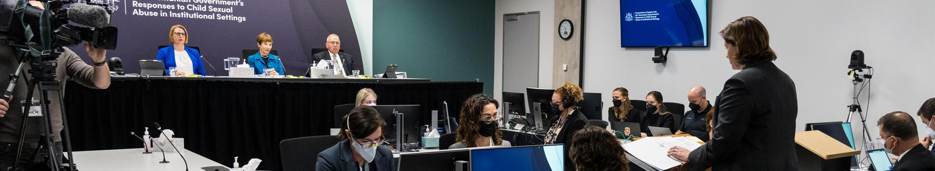 A person with a camera filming a person standing in front of the commissioners, between them are people sitting at desks with computer screens wearing face masks